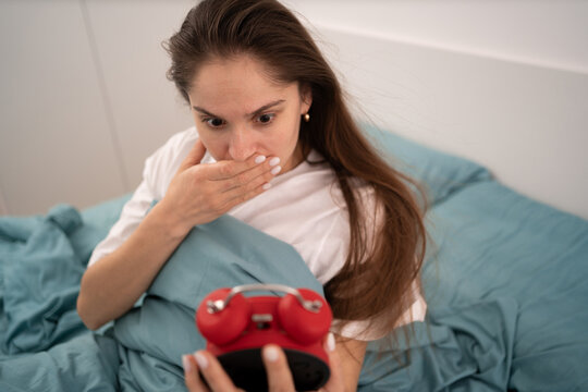 Bad morning concept. Shocked overslept woman waking up at home holding alarm clock lying in bed late for work . Oversleeping Problem.