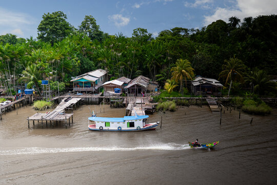 View of traditional wooden stilt houses and dense tropical jungle along the Amazon River with a person in a small motorized boat speeding across the water.