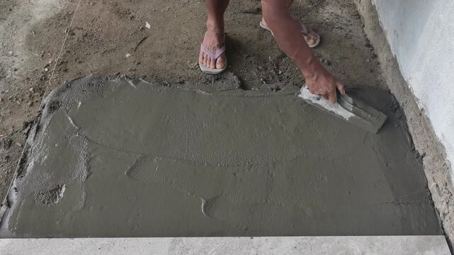 Construction worker smoothing and spreading wet cement mixture on a floor using a trowel. Skilled plastering and floor leveling work.