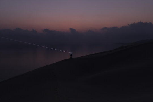 View of a person with a headlamp standing on a dark sand dune overlooking the sea during twilight in Socotra, Yemen.
