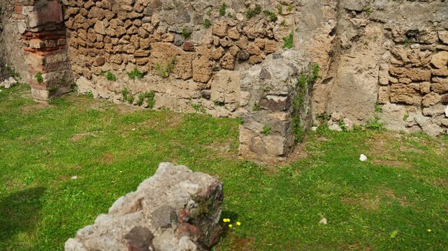 View across a Pompeii room ruin with rough stone walls, surviving masonry sections, and grass floor, showing the excavated remains of an ancient Roman domestic space.