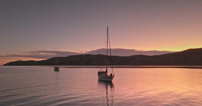 Two sailboats anchoring in serene lagoon at colorful sunset, reflecting tranquil dusk colors across still ocean water with mountains silhouetted on the horizon. Travel, explore tropical Fiji islands