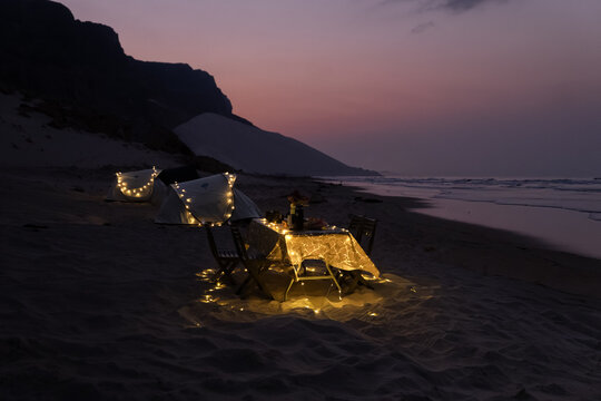 View of a beach campsite with illuminated tents and a dinner table under a purple twilight sky in Socotra, Yemen.