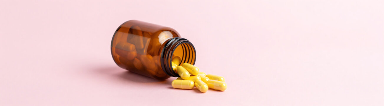 shot open amber supplement bottle releasing cascade yellow capsules onto soft pink pastel background ample