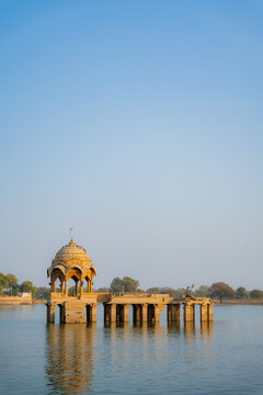 Traditional sandstone pavilion standing in Gadisar Lake with calm water reflections and clear sky in Jaisalmer, Rajasthan, India.