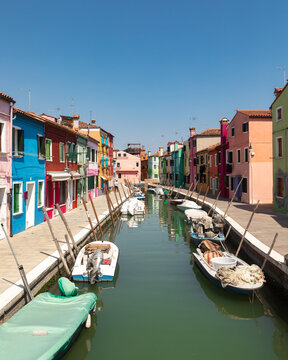 View of colorful houses lining a narrow canal with small boats moored along the stone walkways under a clear blue sky Venice, Veneto, Italy.