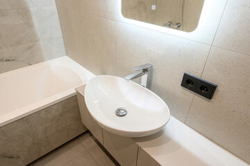 Fototapeta premium modern bathroom with a white oval sink on a sleek countertop, built-in bathtub, illuminated mirror, chrome faucet, neutral marble-textured tiles, dual power outlets.