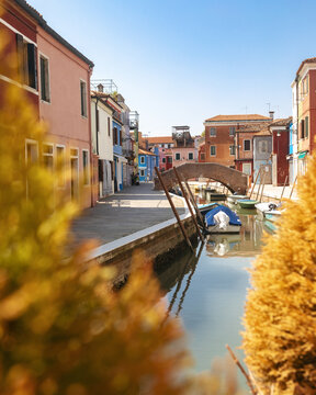 View of the colorful houses and a small stone bridge over a canal with moored boats in Burano, Venice, Veneto, Italy.