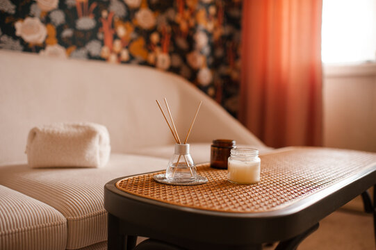 Cozy home atmosphere with liquid home perfume in glass bottle and sticks with candles on rattan table with couch in living room. Hygge domestic interior.