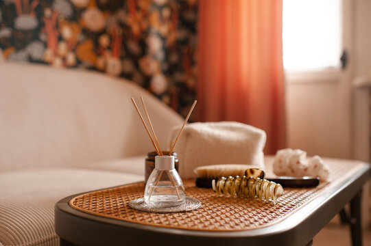 Cozy home atmosphere with liquid home perfume in glass bottle and sticks with candles on rattan table with couch in living room. Hygge domestic interior.