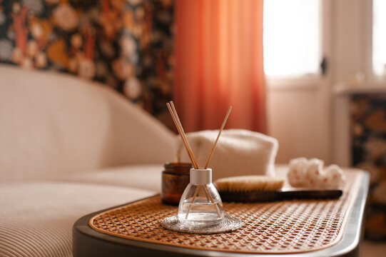 Cozy home atmosphere with liquid home perfume in glass bottle and sticks with candles on rattan table with couch in living room. Hygge domestic interior.