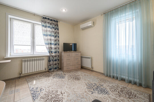 light-filled room with two windows, sheer light blue curtains, patterned rug, wooden dresser with TV, wall-mounted AC, radiators, tiled floor, and geometric-patterned drapes. Cozy and bright
