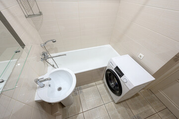 Fototapeta premium compact bathroom with beige tiles, white bathtub, sink on pedestal, toilet, wall-mounted mirror, metal shower rack, washing machine, and grab bar. Clean and functional layout.