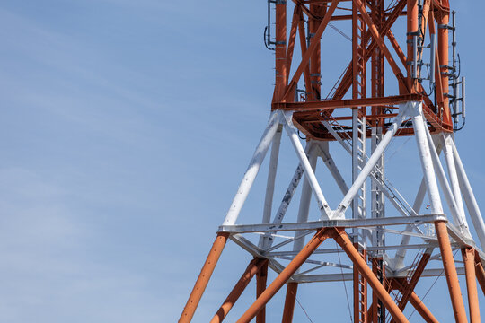 Close-up red-and-white transmission tower under calm blue sky, emphasizing telecommunication industry's infrastructure and showcasing structure's lattice framework, cables, and weathered surface.