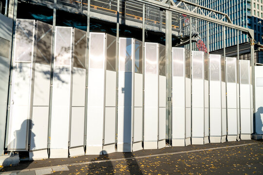 White folding metal construction gate at an urban building site. Perforated panels reflect bright sunlight in a city environment. Temporary fencing with background crane and skyscraper view.