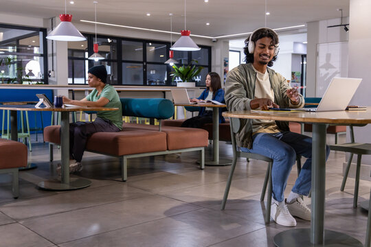 Diverse freelancers with Indian man wearing headphones using laptop in coworking cafe, copy space