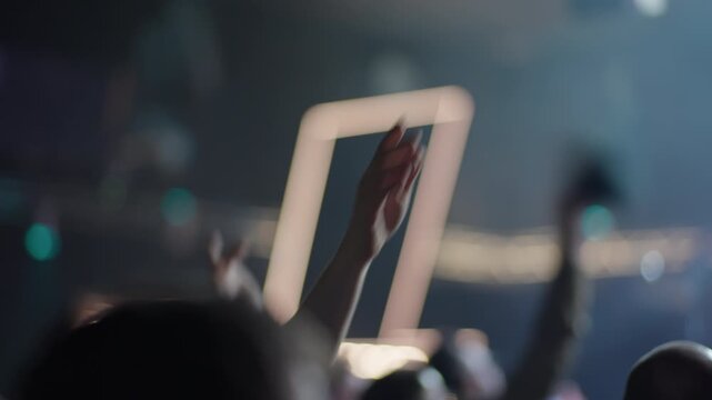Blurred shot of energetic crowd at music festival. People wave hands in air in front of glowing stage lights. Silhouettes of fans cheering dancing at live concert show. Atmospheric vibrant
