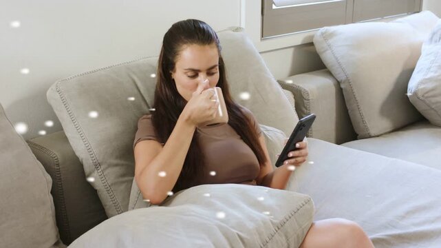 Woman on couch, glancing phone, scrolling and sipping mug, particles floating over woman for ads