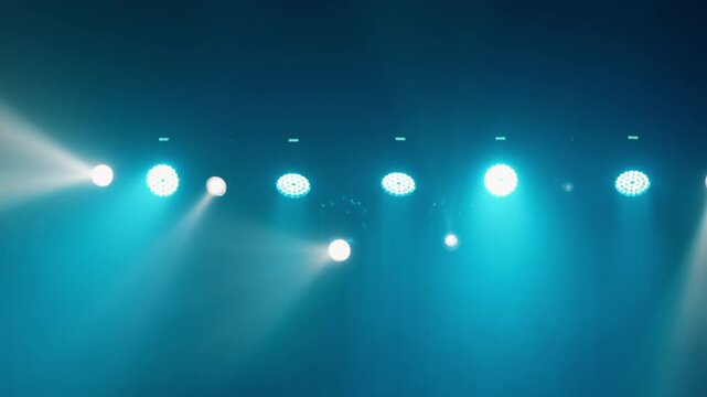Row of cyan teal LED par lights alternating with white spotlights, thick smoke makes beam visible against dark teal background, atmospheric