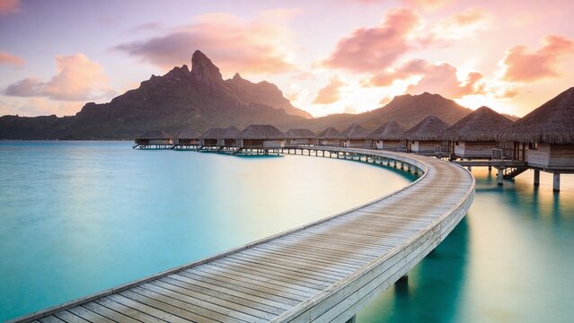 A serene wooden walkway leads to huts on stilts over calm turquoise waters at sunset with majestic mountains in the background near a tropical island.