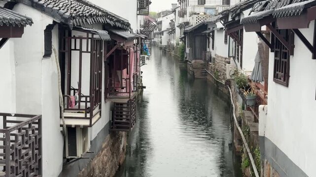 Serene canal flowing between traditional Chinese whitewashed buildings with dark timber lattice windows and overhanging balconies on a rainy day in a historic water town