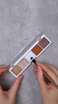A woman's hands test an eyeshadow palette with a brush on the skin of her hands on a table.