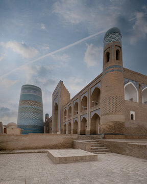 View of the Kalta Minor Minaret and Muhammad Amin-khan Madrasah with intricate blue tilework and brick arches under a cloudy sky in Khiva, Xorazm Region, Uzbekistan.