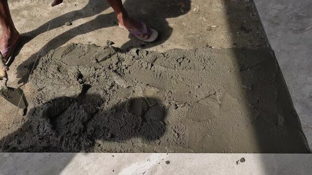 Construction worker leveling and spreading wet cement mix on a floor surface using a trowel. Skilled floor finishing and concrete work.