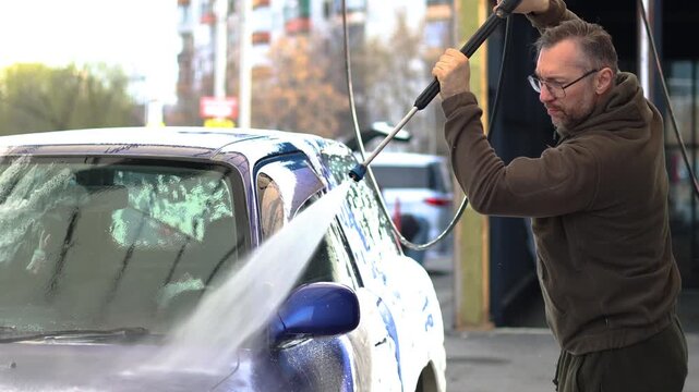 A middle aged man washes a blue car at a self-service car wash. The car wash's high-pressure jet sprays water and rinses away the cleaning foam.