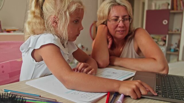 Girl studying with laptop and workbook at desk, mother supporting learning process. Home schooling and parenting support