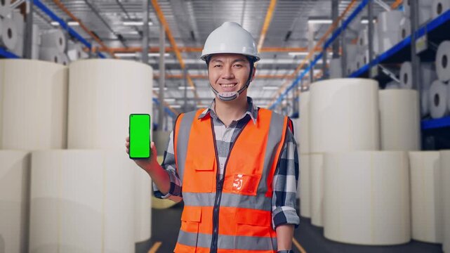 Asian Male Engineer With Safety Helmet Smiling And Showing Green Screen Smartphone To The Camera While Standing at Warehouse with Rows of Stacked White Polymer and Paper Rolls