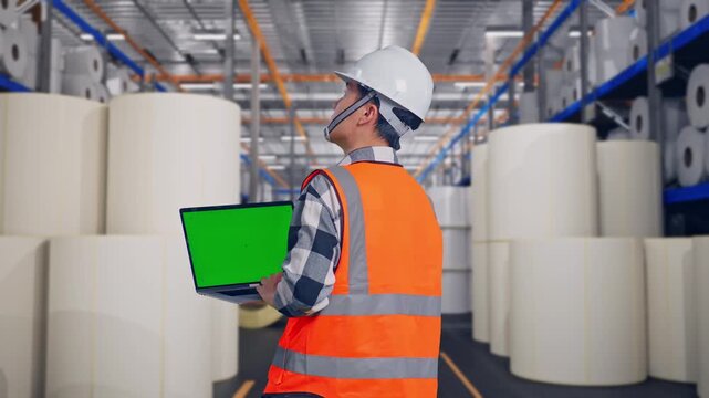 Back View Of Asian Male Engineer With Safety Helmet Working On A Green Screen Laptop And Looking Around While Standing at Warehouse with Rows of Stacked White Polymer and Paper Rolls
