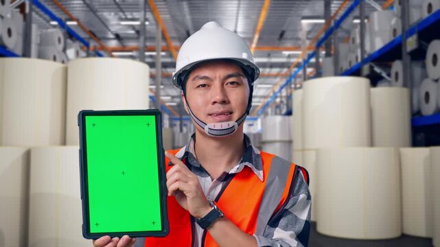 Close Up Of Asian Male Engineer With Safety Helmet Smiling And Showing Green Screen Tablet To The Camera While Standing at Warehouse with Rows of Stacked White Polymer and Paper Rolls