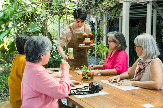 Young cafe server brings iced drink and dessert to senior Asian women meeting again after long separation, starting heartfelt lunch conversation about life change, friendship support, comfort, warmth.
