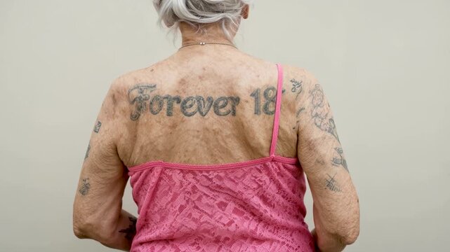 Senior woman with forever eighteen tattoo on her back showing aging process and rebellious spirit of elderly person