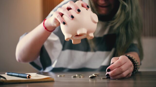 Young broke woman empty piggy bank, shaking it to get last coins out on wooden table. Concept financial problems, poverty, savings and budget planning at home