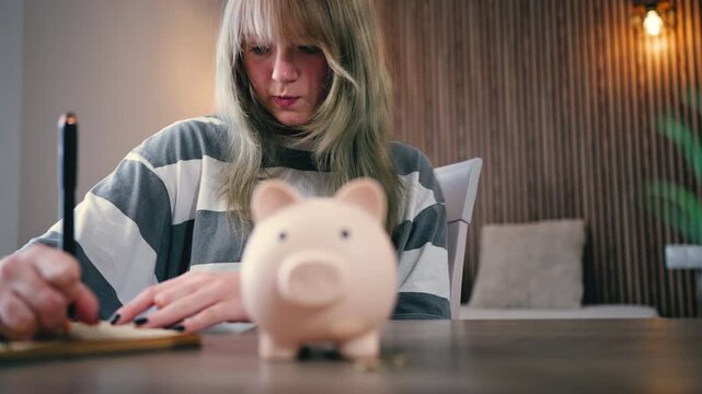 Concentrated teenager girl sit at table meticulously writing down budget calculations, with pink piggy bank in foreground . Concept personal finance and saving money