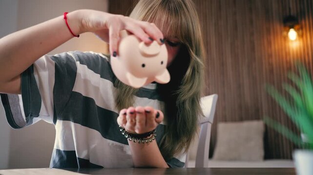 Sad and broke teenage girl desperately trying to get money from empty piggy bank. Concept  struggle with personal finances, poverty, and challenge of saving during recession
