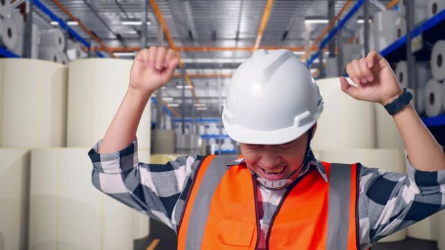 Close Up Of Asian Male Engineer With Safety Helmet Screaming Goal And Dancing Celebrating at Warehouse with Rows of Stacked White Polymer and Paper Rolls