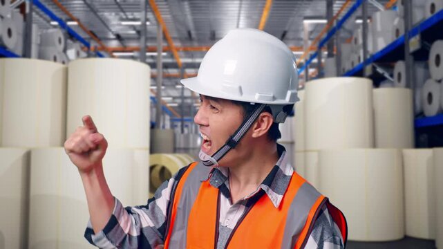 Close Up Side View Of Angry Asian Male Engineer With Safety Helmet Shouting At Someone While Standing at Warehouse with Rows of Stacked White Polymer and Paper Rolls