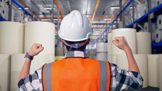 Close Up Back View Of A Male Engineer With Safety Helmet Raising His Hands Celebrating While Working at Warehouse with Rows of Stacked White Polymer and Paper Rolls