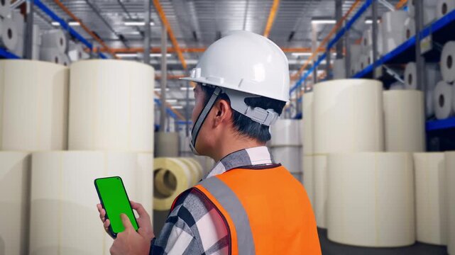 Close Up Back View Of Asian Male Engineer With Safety Helmet Working On A Green Screen Smartphone And Looking Around While Standing at Warehouse with Rows of Stacked White Polymer and Paper Rolls
