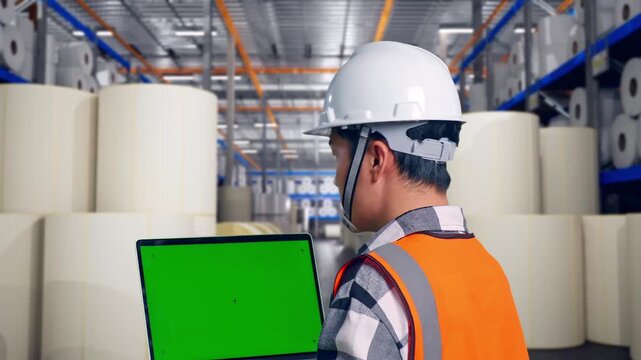 Close Up Back View Of Asian Male Engineer With Safety Helmet Working On A Green Screen Laptop And Looking Around While Standing at Warehouse with Rows of Stacked White Polymer and Paper Rolls