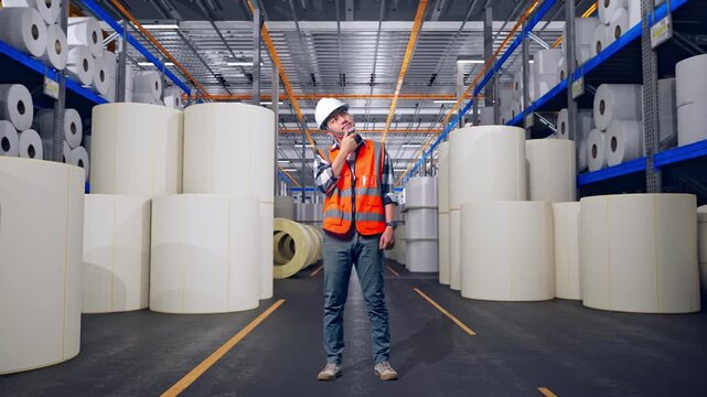 Full Body Of Asian Male Engineer With Safety Helmet Thinking And Looking Around Then Raising His Index Finger While Standing at Warehouse with Rows of Stacked White Polymer and Paper Rolls