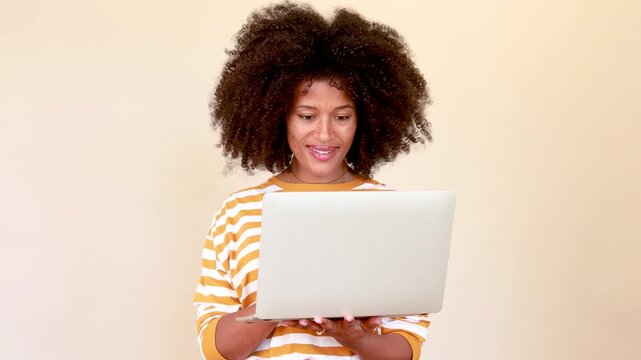 Smiling african american woman with afro hair using laptop for remote work or online communication on a beige background