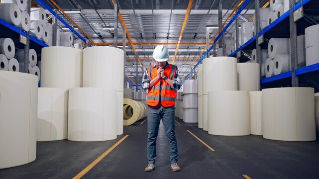 Full Body Of Asian Male Engineer With Safety Helmet Pray For Something While Standing at Warehouse with Rows of Stacked White Polymer and Paper Rolls