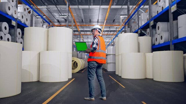 Full Body Back View Of Asian Male Engineer With Safety Helmet Working On A Green Screen Laptop And Looking Around While Standing at Warehouse with Rows of Stacked White Polymer and Paper Rolls