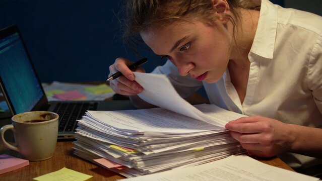 Young woman working late at office desk sorting documents paperwork stack with focused business workflow, overtime job stress organization and professional data management in modern workplace