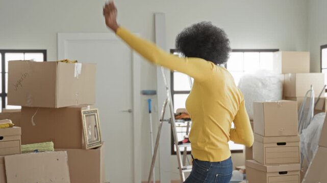 Woman raising arms dancing beside stacked cardboard boxes inside a building during a move; celebration joy.