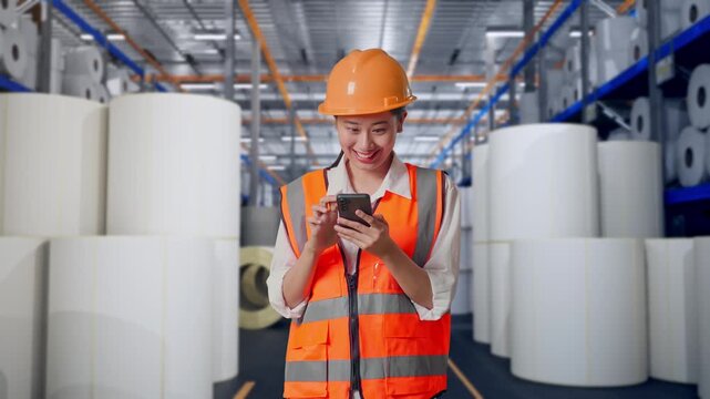Asian Female Engineer With Safety Helmet Using Smartphone While Standing at Warehouse with Rows of Stacked White Polymer and Paper Rolls